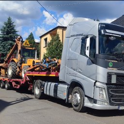 PTUH Jan Cieślik - Laweta Volvo FH 500 przewozi pomarańczową koparko-ładowarkę Renault na czerwonej platformie. Widok z boku na tle zabudowań i błękitnego nieba.