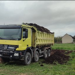 PTUH Jan Cieślik - Żółta wywrotka Mercedes Actros z pełną skrzynią ziemi na tle zielonej łąki i zabudowań. Widoczne ślady opon na ziemi. Pochmurne niebo.