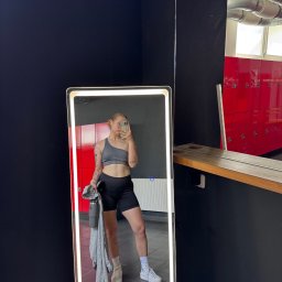 Zuzanna Śliwińska - Osoba w sportowym stroju pozuje do selfie w lustrze na tle czerwonych szafek w szatni. W ręku trzyma butelkę wody i bluzę. Widoczne tatuaże na ciele.