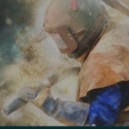 SPAW-MET - Pracownik w kombinezonie i masce ochronnej piaskuje metalową konstrukcję. Wokół unoszą się drobinki piasku, oświetlone jasnym światłem.