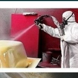 SPAW-MET - Pracownik w kombinezonie ochronnym maluje natryskowo element w kolorze kremowym w warsztacie. Widoczny pistolet natryskowy i czerwona ściana w tle.