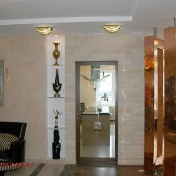 KWW-REMART Remigiusz Zagrodnik - Eleganckie wnętrze salonu z beżowymi ścianami, czarną sofą z poduszkami w paski, dekoracyjnymi niszami z oświetleniem i ozdobami, oraz miedzianą ścianką działową z otworami, widok na kuchnię przez...