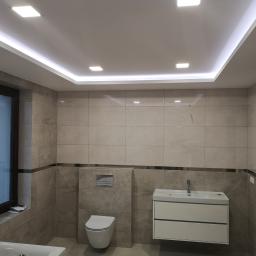 KWW-REMART Remigiusz Zagrodnik - Wykończona łazienka w beżowych płytkach z podwieszaną toaletą, białą szafką umywalkową i oknem; widoczne oświetlenie sufitowe LED i fragment wanny.