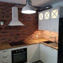 KWW-REMART Remigiusz Zagrodnik - Nowoczesna kuchnia z ceglaną ścianą, białymi szafkami z podświetleniem, drewnianym blatem i czarnym zlewem oraz piekarnikiem.