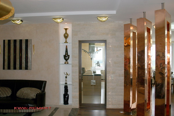 Eleganckie wnętrze salonu z beżowymi ścianami, czarną sofą z poduszkami w paski, dekoracyjnymi niszami z oświetleniem i ozdobami, oraz miedzianą ścianką działową z otworami, widok na kuchnię przez...