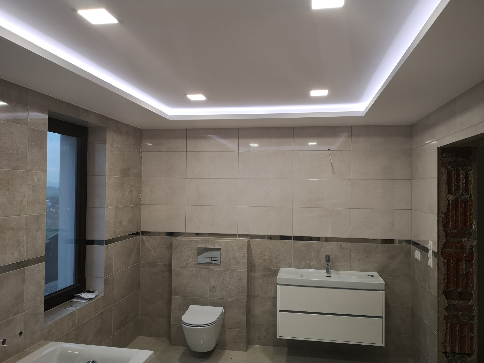 Wykończona łazienka w beżowych płytkach z podwieszaną toaletą, białą szafką umywalkową i oknem; widoczne oświetlenie sufitowe LED i fragment wanny.