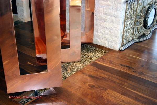 Fragment wnętrza z drewnianą podłogą w jodełkę, dekoracyjnym panelem z miedzianymi ramami i żwirowym wypełnieniem oraz fragmentem kamiennej ściany z ozdobnym elementem.