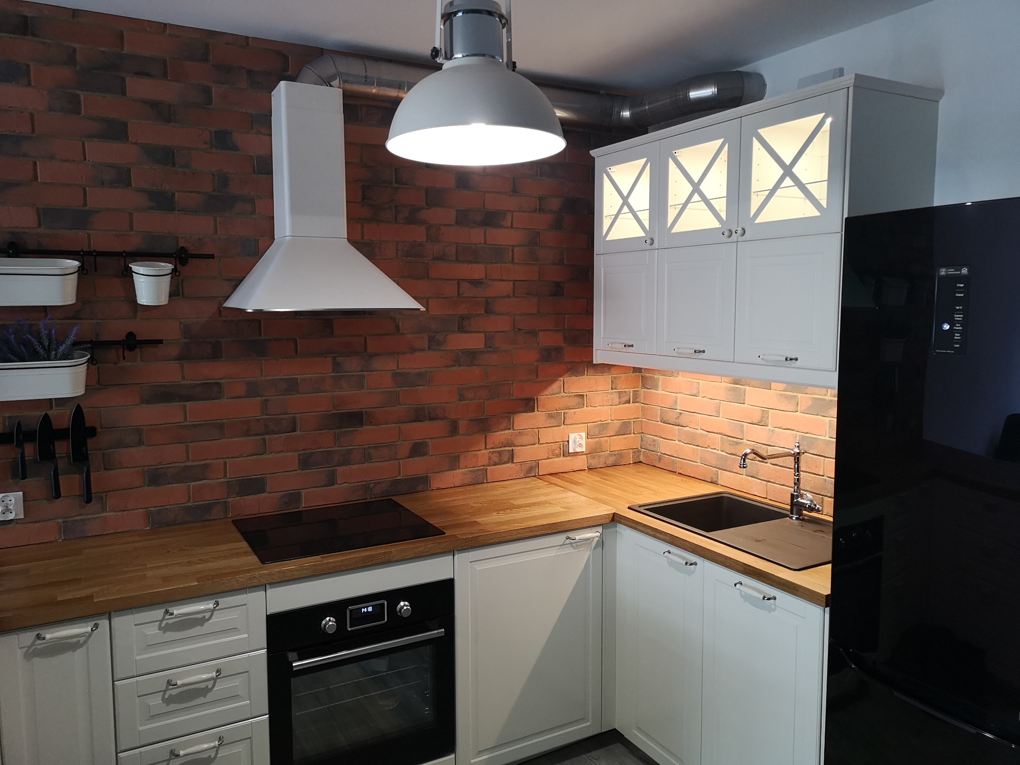 Nowoczesna kuchnia z ceglaną ścianą, białymi szafkami z podświetleniem, drewnianym blatem i czarnym zlewem oraz piekarnikiem.