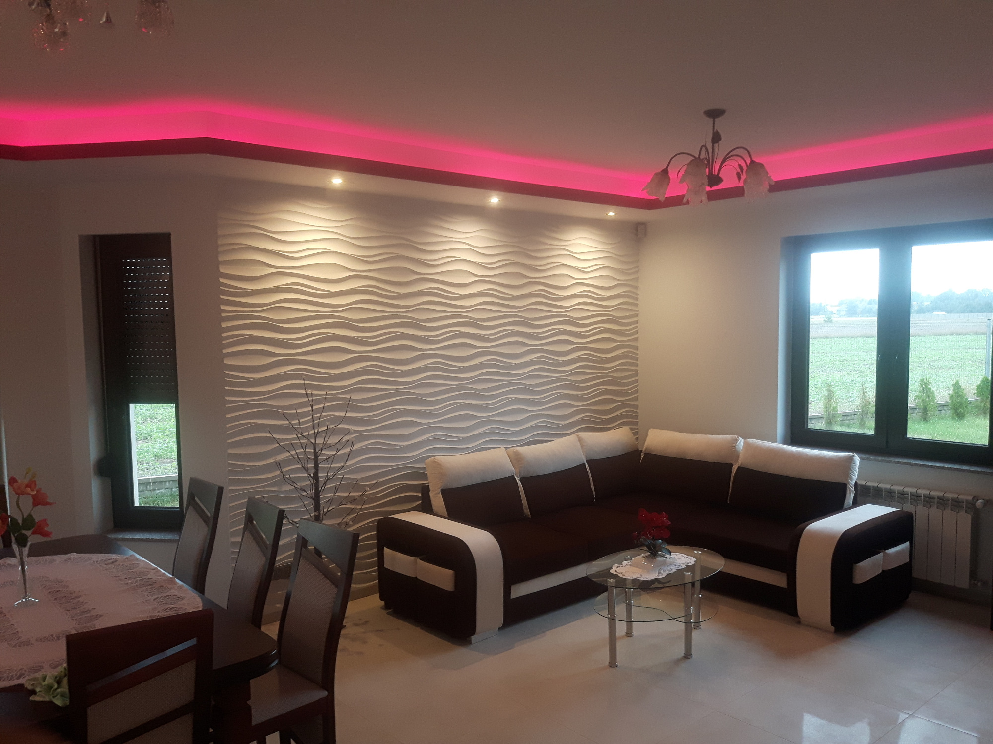 Salon w Bralinie z dekoracyjną ścianą 3D w kształcie fal, podświetleniem LED w kolorze różowym, brązowym narożnikiem i widokiem na zieleń za oknem.