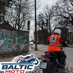 OSK Baltic Moto - Instruktor na motocyklu w kamizelce z literą 'L' na brukowanej ulicy, widok z tyłu, w tle budynki i drzewa bez liści, pochmurne niebo.