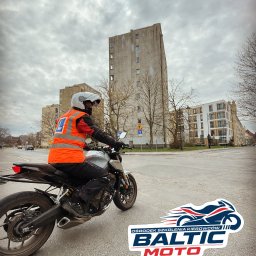 OSK Baltic Moto - Instruktor w kamizelce na motocyklu na skrzyżowaniu, w tle budynki i pochmurne niebo. Na asfalcie naklejka z logo ośrodka szkolenia kierowców.