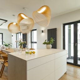 Villa.M - Jasne wnętrze apartamentu z wyspą kuchenną, drewnianymi krzesłami i designerskimi lampami. Widok na balkon i część salonu z sofą. Nowoczesny design.