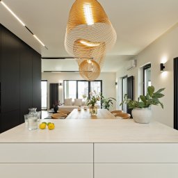 Villa.M - Nowoczesne wnętrze: biała wyspa kuchenna z cytrynami, czarne szafki, designerskie lampy i widok na salon z sofą. Minimalistyczny, jasny wystrój.