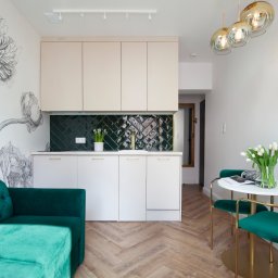 KMK Meble - Kompaktowa kuchnia z białymi frontami i ciemnozieloną glazurą w jodełkę. Uwagę przykuwa fototapeta z motywem kwiatowym i szmaragdowa sofa. Całość dopełnia parkiet w jodełkę.