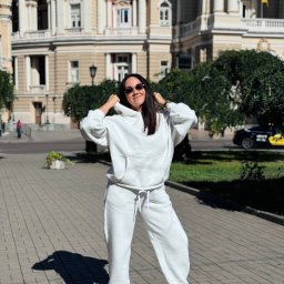 Szwalnia Andrei Jilnicov - Uśmiechnięta brunetka w białym dresie z kapturem, pozuje na tle budynku opery z flagą Ukrainy. Dres uszyty na miarę, z dbałością o detale i komfort noszenia.