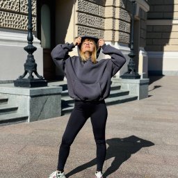 Szwalnia Andrei Jilnicov - Kobieta w szarym, obszernym hoodie z kapturem, czarnych legginsach i sportowych butach pozuje na tle budynku z dekoracyjnymi detalami architektonicznymi.
