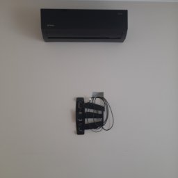 Łukasz Sordyl - Czarny klimatyzator na ścianie nad uchwytem z kablami. Widoczne przewody. Minimalistyczny design urządzenia.