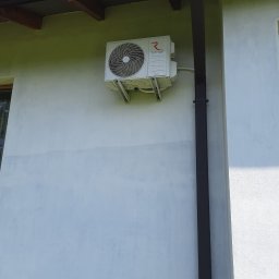 Łukasz Sordyl - Biała jednostka zewnętrzna klimatyzacji Rotenso ELIS zamontowana na ścianie budynku, obok brązowej rynny spustowej. Widoczne elementy elewacji i fragment nieba.
