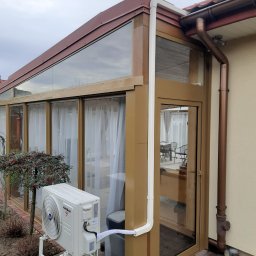 Łukasz Sordyl - Klimatyzator Kaisai zamontowany na zewnątrz oranżerii, z widocznymi rurami odprowadzającymi. Na pierwszym planie krzew z czerwonymi owocami. Pochmurne niebo w tle.