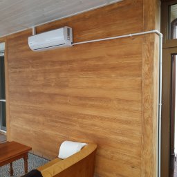 Łukasz Sordyl - Biały klimatyzator na ścianie z drewnianą okładziną. Widoczne rury instalacyjne. Wnętrze z sofą i stolikiem kawowym. Jasne pomieszczenie.