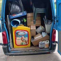 Andrzej Bluszcz - Niebieski bus z otwartymi drzwiami, wypełniony różnymi przedmiotami: kartony, krzesła, pojemniki, reklamy Pepsi i Koral, sugerujące opróżnianie mieszkania lub transport.