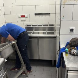 Andrzej Bluszcz - Pracownik w niebieskiej koszulce demontuje metalowy zlew w pomieszczeniu z białymi kafelkami. Na blacie obok leżą narzędzia i akcesoria.