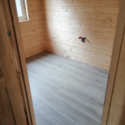 FHU REM-BUD Krzysztof Jastrzębski - Wnętrze domu szkieletowego z drewnianą boazerią i szarą podłogą. Widoczne okno i instalacja wodna. Ujęcie zza drewnianej futryny.