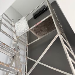FROZE Klimatyzacja i Wentylacja - Czarna jednostka klimatyzacji na szarej ścianie, widziana z dołu przez metalowe rusztowanie. Ujęcie z perspektywy montera podczas instalacji.