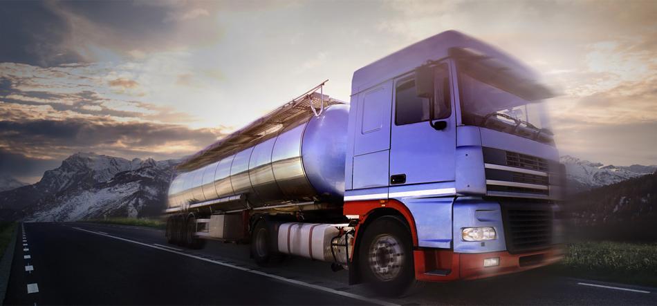 Ciężarówka cysterna w kolorze niebieskim, jadąca asfaltową drogą z ośnieżonymi górami w tle pod pochmurnym niebem.