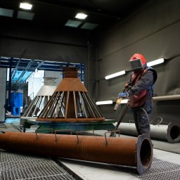 Dozamech - Pracownik w kombinezonie ochronnym przygotowuje metalowe elementy do obróbki w hali. Widoczne detale konstrukcji i narzędzia.