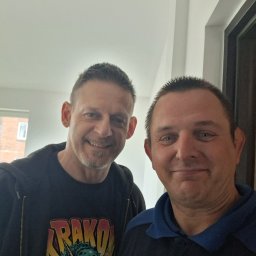 infobud - Uśmiechnięci mężczyźni w pomieszczeniu z oknem w tle. Jeden w bluzie z nadrukiem, drugi w koszulce polo. Zdjęcie z bliska, dobre oświetlenie.