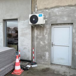 PRIMACOOL - Biała jednostka zewnętrzna klimatyzacji Midea zamontowana na szarej, surowej ścianie budynku obok białych drzwi i okna. Widoczne słupki i pachołek ostrzegawczy.
