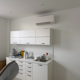 PRIMACOOL - Białe wnętrze gabinetu z klimatyzatorem Daikin na ścianie, nad szafkami. Widoczny fotel dentystyczny i umywalka. Minimalistyczny design, jasne oświetlenie.