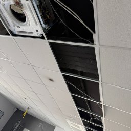 PRIMACOOL - Montaż klimatyzacji kasetonowej w biurze w Kamieniu Pomorskim. Widoczne otwarte panele sufitowe i instalacja urządzenia. Ujęcie z dołu, pod kątem.