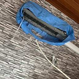 PRIMACOOL - Klimatyzator w niebieskim pokrowcu ochronnym podczas serwisu, widoczne przewody i panel sterowania na tle ściany z imitacji kamienia.