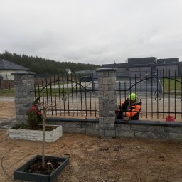 Architektura Ogrodowa - Montaż metalowego ogrodzenia z kamiennymi słupkami. Pracownik w odblaskowej kamizelce przy pracy. W tle domy i drzewa. Na pierwszym planie posadzone drzewka.