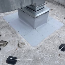 RB System - Szczelne połączenie komina z dachem pokrytym papą, zabezpieczone szarą powłoką. Widoczne elementy mocujące i uszczelniające. Dach pokryty drobnymi zabrudzeniami.