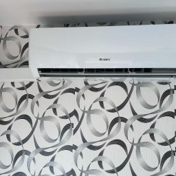 KLIMA STYL SYSTEMS Maciej Sobstyl - Biały klimatyzator GREE zamontowany na ścianie z wzorzystą tapetą w odcieniach szarości i bieli. Widoczny fragment sufitu i ściany bocznej.