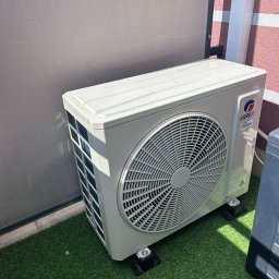 KLIMA STYL SYSTEMS Maciej Sobstyl - Zamontowana jednostka zewnętrzna klimatyzacji Gree na sztucznej trawie, widoczna z boku. Szara ściana w tle, fragment różowej ściany po prawej.