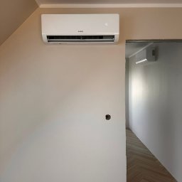 SB Elektro - Biały klimatyzator KaiSai na beżowej ścianie w nowym wnętrzu. Widoczne gniazdka elektryczne i fragment podłogi z parkietem. W tle widoczny drugi klimatyzator.