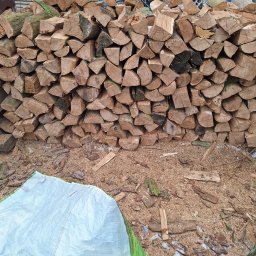 Usługi ogrodnicze Anna Kajda - Ułożone drewno kominkowe w Krapkowicach, przygotowane do sezonu grzewczego. Widoczne kawałki drewna i trociny. Częściowo zakryte plandeką.