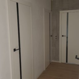 DKFORMA - Białe skrzydła z czarnymi akcentami i klamkami w korytarzu z drewnianą podłogą. Widoczne trzy sztuki, w tym jedno z szarą ścianą w tle.