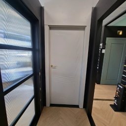 DKFORMA - Jasne, dwuskrzydłowe drzwi z delikatnym frezowaniem, prezentowane w salonie z podłogą ułożoną w jodełkę i fragmentem drzwi szklanych po lewej stronie.