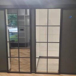 DKFORMA - Nowoczesne, dwuskrzydłowe drzwi przesuwne z czarnymi szprosami, oddzielające pomieszczenia. Widoczny fragment podłogi i ściany w odcieniach szarości.