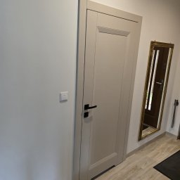 DKFORMA - Zamknięte, beżowe drzwi wewnętrzne z czarną klamką, widoczne z boku na tle białej ściany i drewnopodobnej podłogi. W tle lustro w złotej ramie.