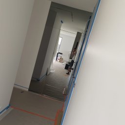 DKFORMA - Wnętrze w trakcie wykańczania: widoczne ściany, podłoga z taśmą malarską i zamontowane lustrzane drzwi. W tle widoczna osoba robiąca zdjęcie.