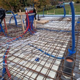 WoodOn Sp. z o.o. - Instalacja ogrzewania podłogowego w domu szkieletowym: rury grzewcze ułożone na izolacji, zbrojenie stalowe, kolektor z rurami czerwonymi i niebieskimi. Etap budowy.