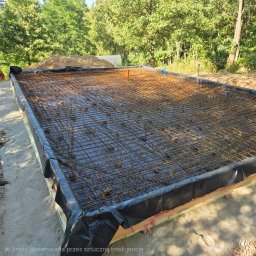 WoodOn Sp. z o.o. - Zbrojenie pod płytę fundamentową z rdzawą siatką zbrojeniową, czarną folią izolacyjną i wystającymi rurami, przygotowane na piaszczystym gruncie w otoczeniu drzew.