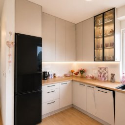 Meble SH składnie mebli - Jasna kuchnia w kształcie litery L z drewnianym blatem, czarną lodówką i przeszkloną szafką z podświetleniem. Minimalistyczny design, ciepłe oświetlenie.