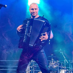 Robert Prusiński - Zespół ,DJ & Akordeon Live - Akordeonista na scenie w niebieskim oświetleniu, z perkusją w tle. Uśmiechnięty muzyk w czarnym stroju gra na akordeonie marki Pigini.